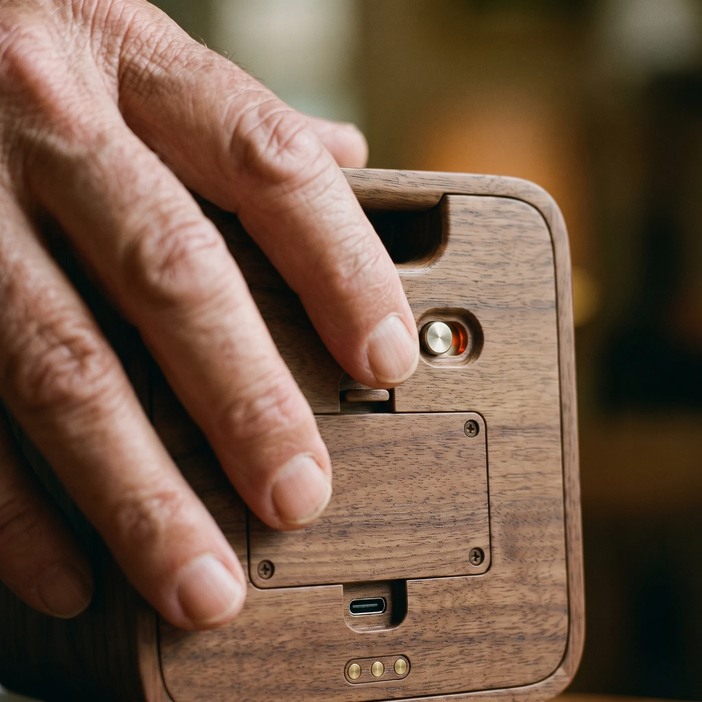 An older hand reaching for the small metal power switch on the back of Roomie, beside the service plate and the USB-C socket.